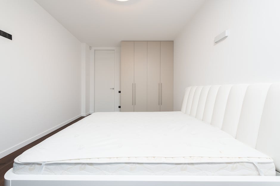 A modern bedroom featuring a large bed with a white mattress and a grey upholstered headboard. The room has floor-to-ceiling windows with sheer white curtains and beige drapes, allowing natural light 