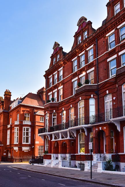 View of a red-brick residential building with multiple large windows, some with white window frames and others with black railings on small balconies. The building's facade is detailed with decorative